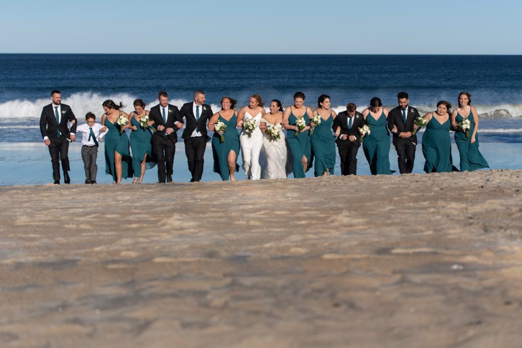 a large group of bridesmaids and bridegrooms walk up the beach with two brides